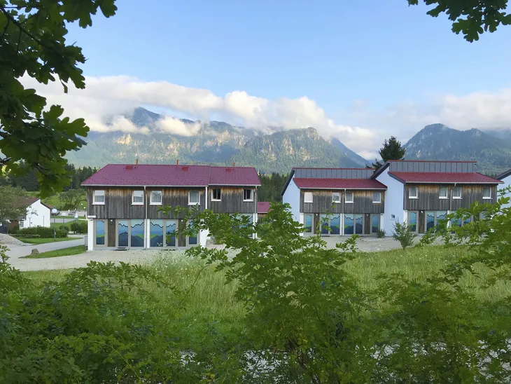Drei Holzhäuser vor Bergkulisse mit blauem Himmel und Wolken.