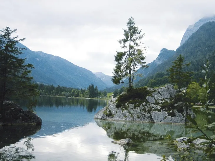 Bergsee mit Bäumen und Felsen, umgeben von bewaldeten Bergen.
