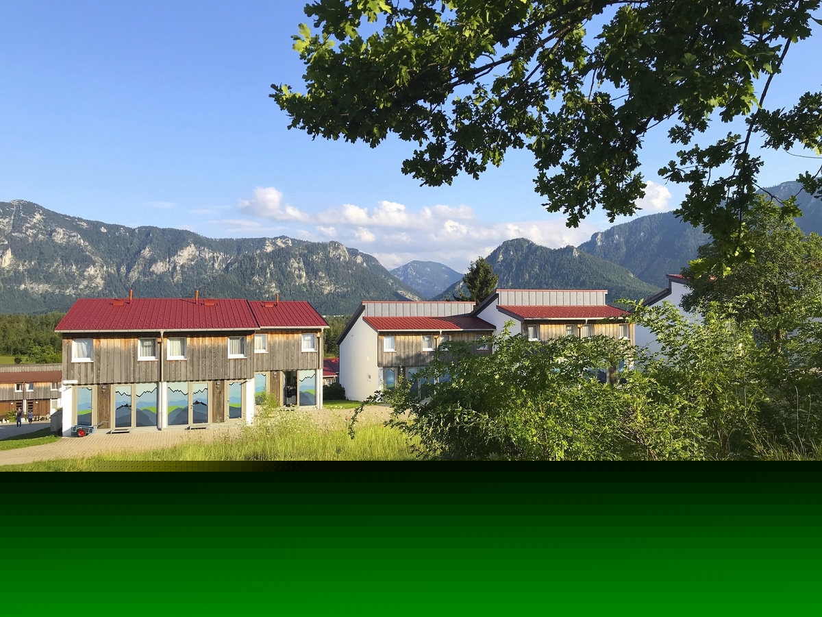 Gebäude vor Berglandschaft mit klarem Himmel und Bäumen im Vordergrund.