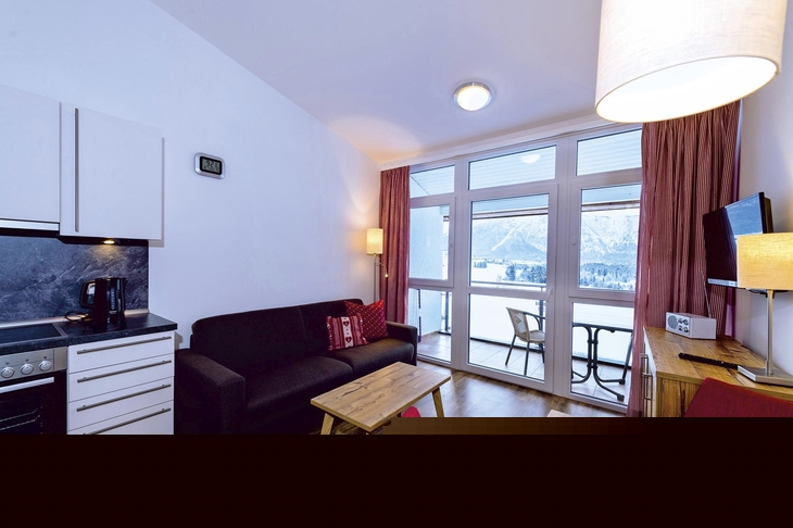 Wohnzimmer mit Küche in Ferienwohnung, Holzboden, Sofa und Fenster mit Bergblick.