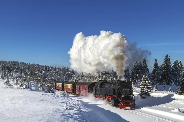 Dampflok fährt durch verschneite Landschaft mit Nadelbäumen unter klarem, blauem Himmel.