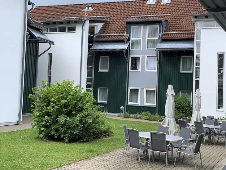 Mehrstöckiges Gebäude mit Terrasse mit Tischen, Stühlen und Sonnenschirmen und grünem Busch auf einer Wiese.