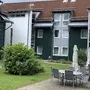 Mehrstöckiges Gebäude mit Terrasse mit Tischen, Stühlen und Sonnenschirmen und grünem Busch auf einer Wiese.