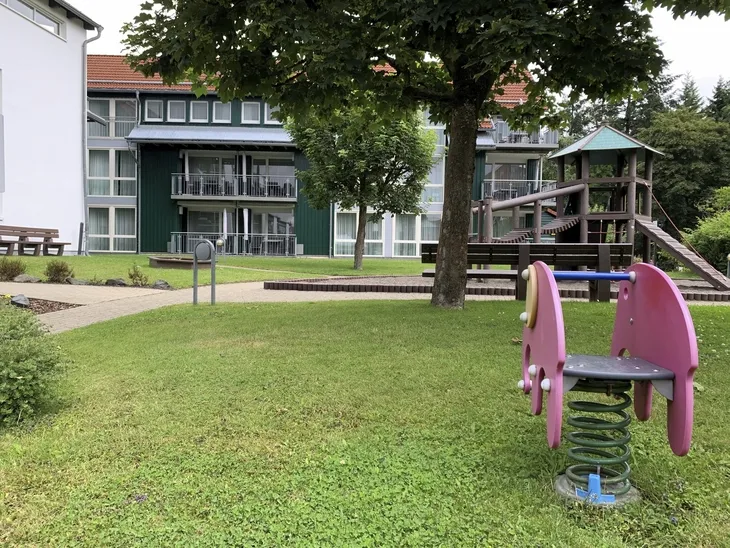 Spielplatz mit Wipptier und Spielturm auf einer Wiese vor einem Gebäude der Ferienanlage Braunlage.