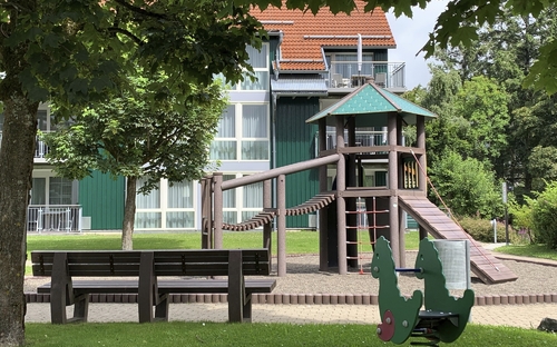 Spielplatz mit Spielturm und Wipptier sowie einer Holzbank vor einem Gebäude der Ferienanlage Braunlage.