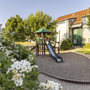 Spielplatz mit Rutsche in einer Gartenanlage vor einem Gebäude am sonnigen Tag.