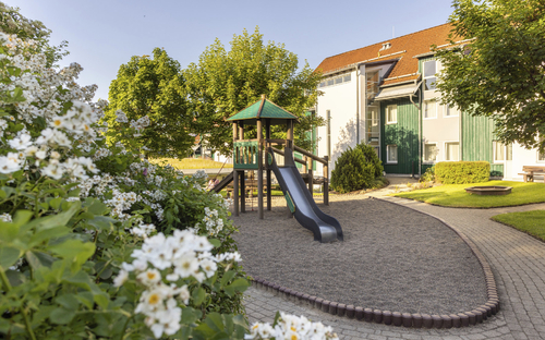 Spielplatz mit Rutsche in einer Gartenanlage vor einem Gebäude am sonnigen Tag.