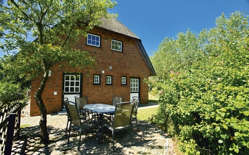 Reedgedecktes Backsteinhaus mit Tisch und Stühlen davor, bei blauem Himmel umgeben von Bäumen, in der Ferienanlage List auf Sylt.