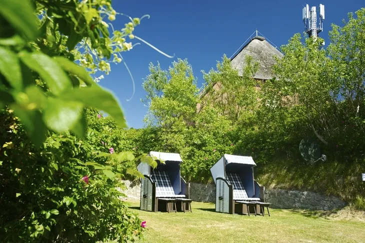 Zwei Strandkörbe auf einer grünen Wiese, von Bäumen umgeben und blauem Himmel, vor einem Backsteinhaus mit Reetdach.