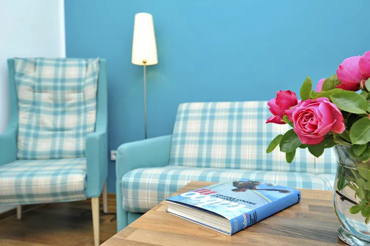 Wohnzimmer mit kariertem Sessel und Sofa, Tisch mit Buch und Vase mit Rosen.