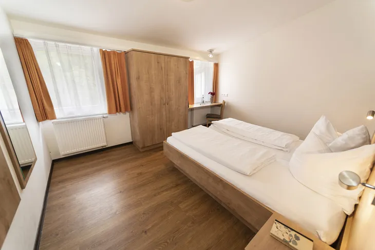 Schlafzimmer mit Doppelbett, Schrank, Fenster mit Vorhängen und Holzboden.