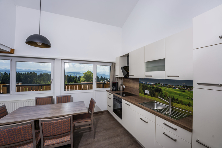 Moderne Küche mit Esstisch, Fenstern und Waldlandschaft im Hintergrund.