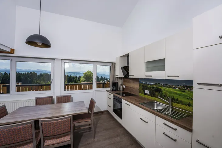 Moderne Küche mit Esstisch, Fenstern und Waldlandschaft im Hintergrund.