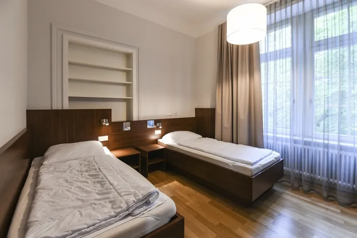 Zwei Einzelbetten in einem hellen Zimmer mit Holzmöbeln und großen Fenstern.