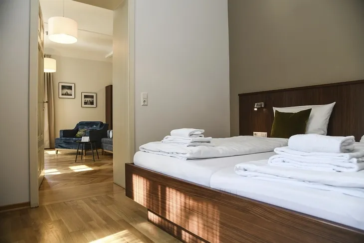 Hotelzimmer mit Bett, weißer Bettwäsche, Holzmöbeln und Blick in ein Wohnzimmer mit Sofa.