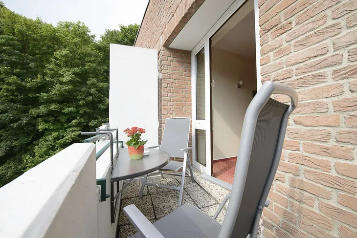 Balkon mit Tisch, zwei Stühlen und einer Pflanze, Ausblick auf Bäume.