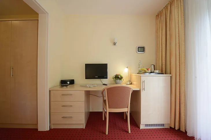 Schreibtisch mit Computer, Lampe, Stuhl und Minibar in einem Hotelzimmer.