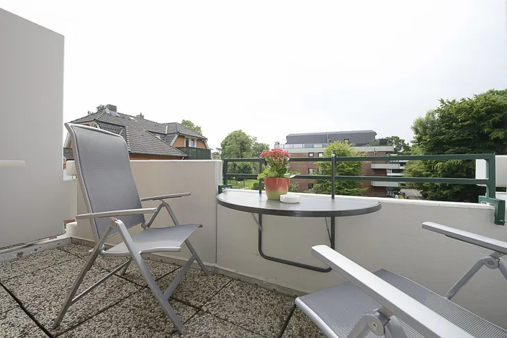 Balkon mit Tisch, Stühlen und Topfpflanze, umgeben von Gebäuden und Bäumen.