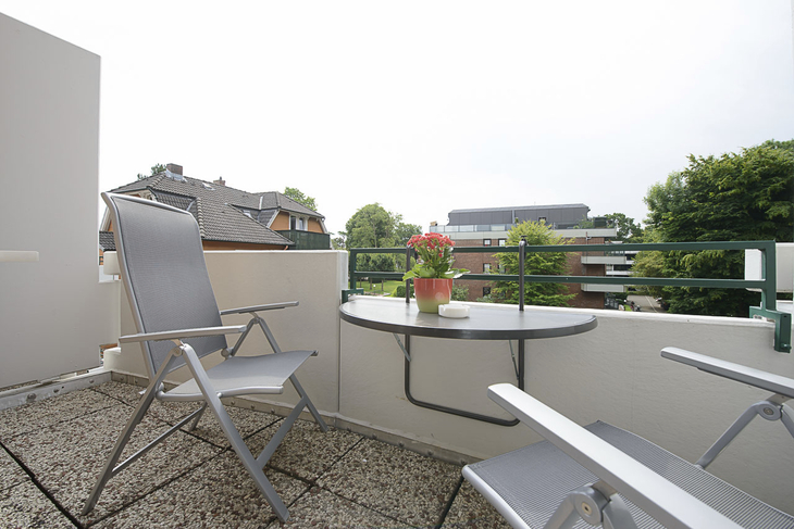 Balkon mit Tisch, Stühlen und Topfpflanze, umgeben von Gebäuden und Bäumen.