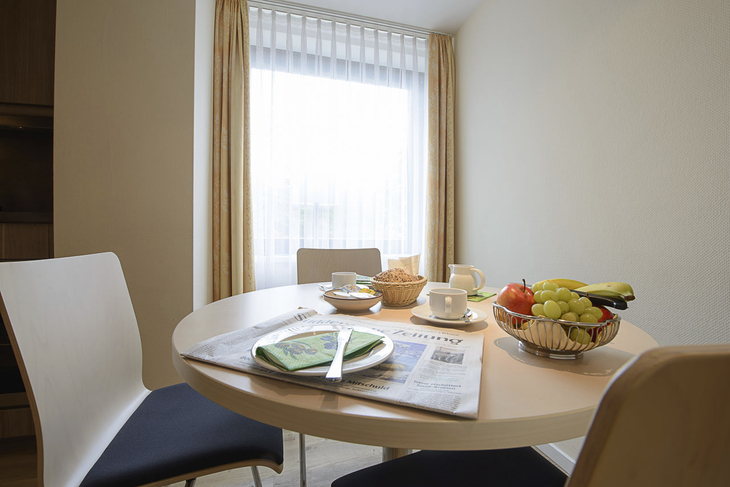 Frühstückstisch mit Obstkorb, Zeitung, Geschirr und heller Fensteransicht.