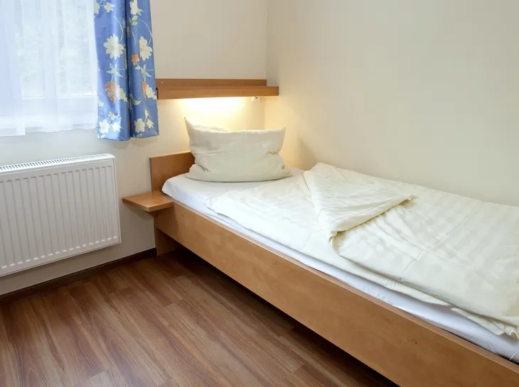 Einfach eingerichtetes Einzelzimmer mit Bett, Bettwäsche und blau gemusterten Vorhängen in der Ferienanlage Prüm