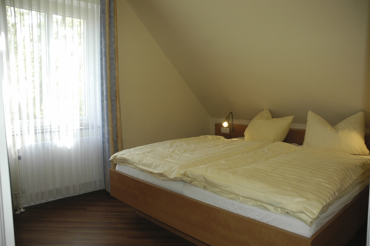 Ein Schlafzimmer mit Doppelbett und heller Bettwäsche, Fenster mit Gardinen links.