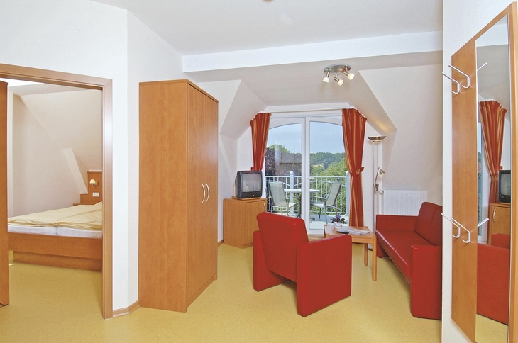 Wohnzimmer in der Ferienanlage Wyk auf Föhr, mit rotem Sofa, Sessel und Fernseher sowie großen Balkontüren mit bunten Vorhängen und Sitzmöbeln auf dem Balkon. Rechts im Bild eine Wandgarderobe mit Spiegel und links im Bild der Blick ins Schlafzimmer.