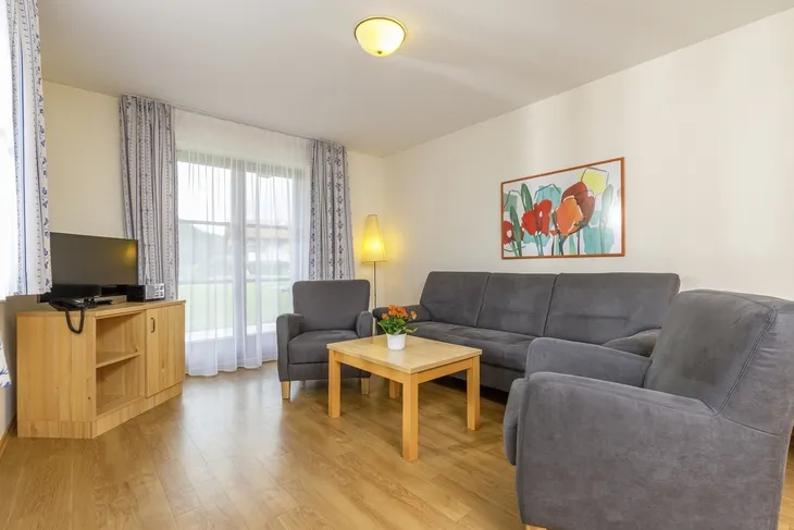 Wohnzimmer mit grauem Sofa, Sesseln, Holztisch und Fernseher.
