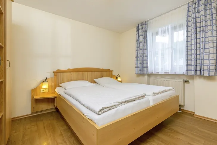 Holz-Doppelbett in hellem Schlafzimmer mit Fenster und blau-weiß karierten Vorhängen.
