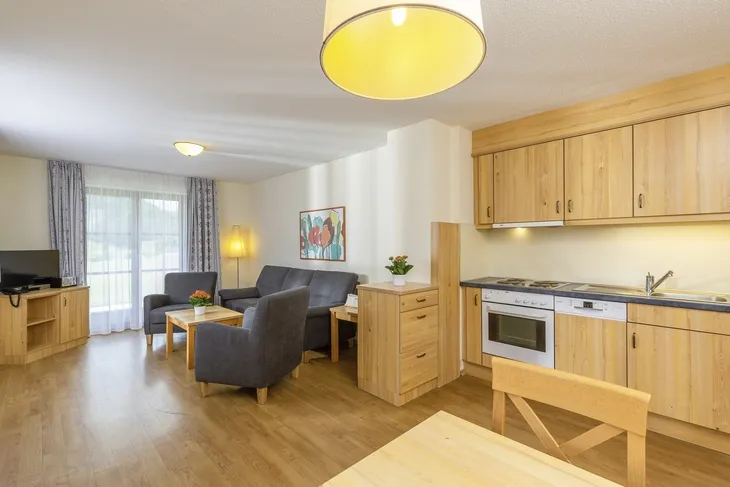 Modernes Wohnzimmer mit Küchenzeile, Sofa, Tisch und Fernseher in hellem Holzdekor.