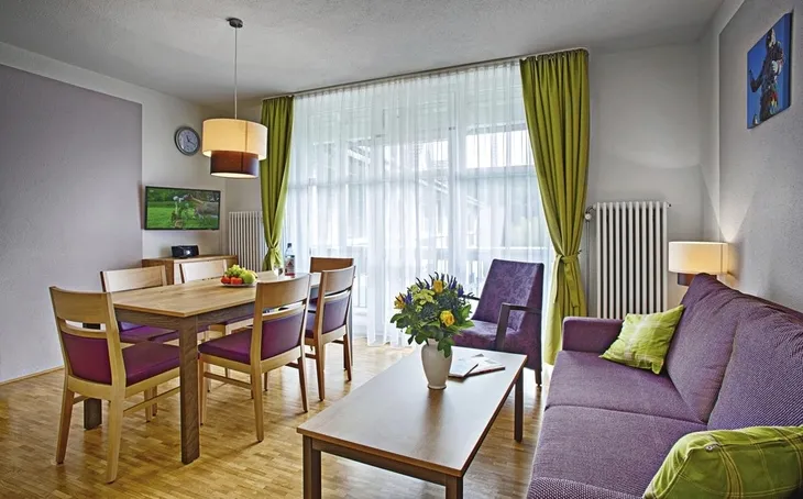 Wohnzimmer mit Esstisch, Sofa, TV, Glasvase mit Blumen, hell durchfenstert.