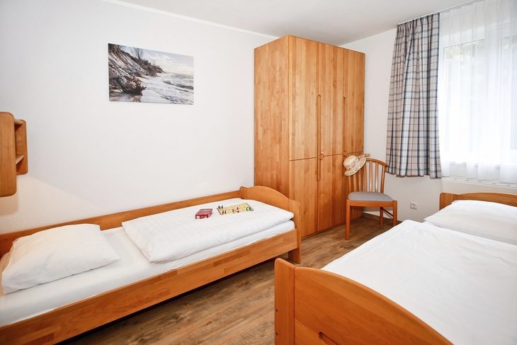 Zwei Einzelbetten in einem hellen Schlafzimmer mit Holzschränken und Fenster mit Vorhängen.