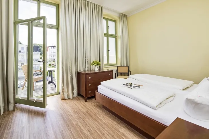 Helles Schlafzimmer mit Doppelbett, Kommode, Balkon mit Gartenmöbeln und Vorhängen.