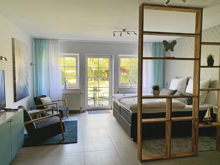 Modernes Einzimmer-Apartment mit Bett, Sesseln, TV und Regalelement, lichtdurchflutet.