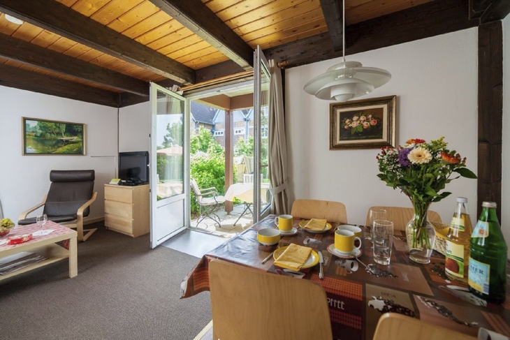 Wohnzimmer mit Essbereich, Holzmöbeln, Blumenstrauß auf Tisch, Blick auf Terrasse.