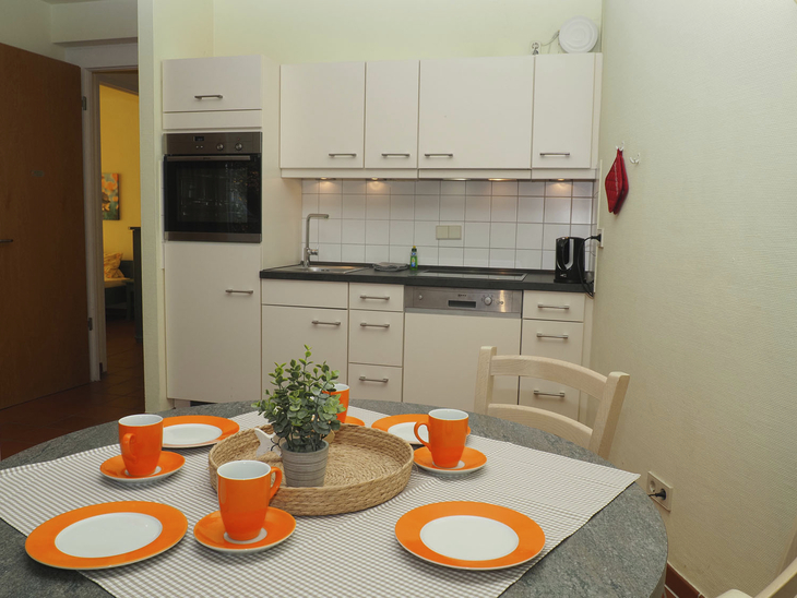 Kleine Küche mit Esstisch, weißem Geschirr und orangefarbenen Tassen.