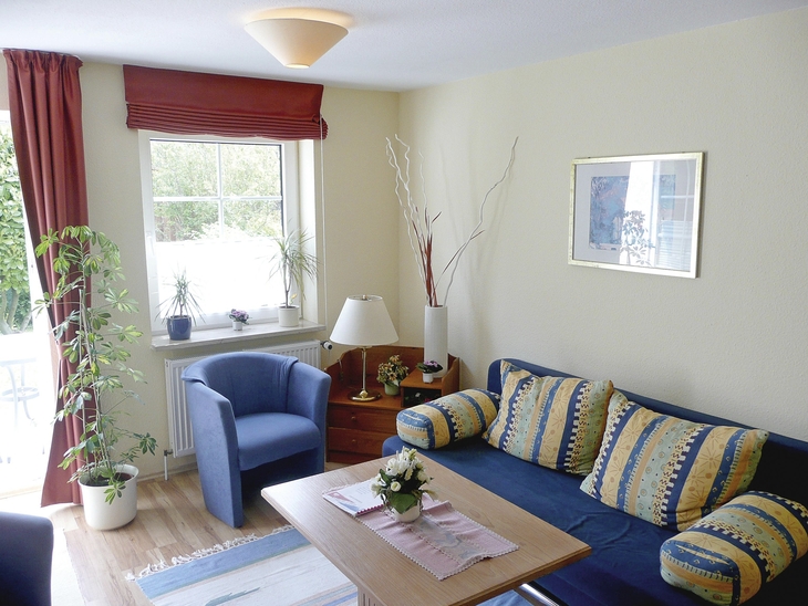 Wohnzimmer mit Sofa, Sessel, Tisch, Pflanzen und einem Fenster mit Vorhängen.
