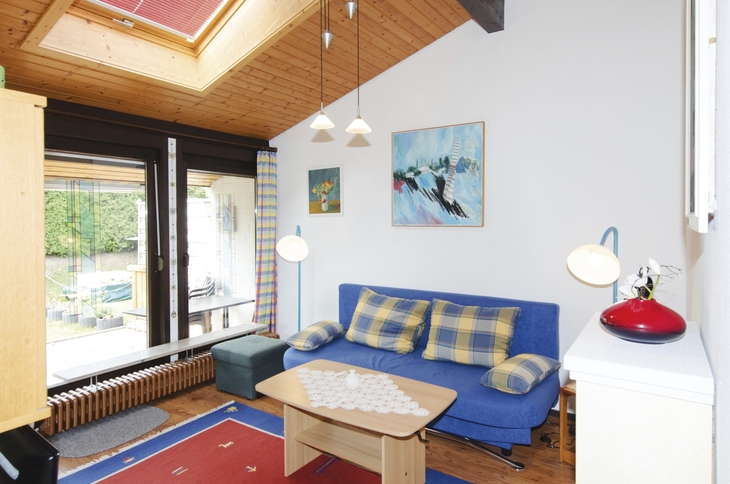 Wohnzimmer mit blauem Sofa, Holztisch, Fenstern und Gemälden an der Wand.