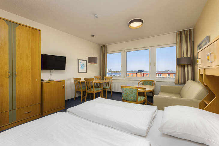 Helles Hotelzimmer mit Doppelbett, Esstisch, Sofa und Fenster mit Blick auf Yachthafen.