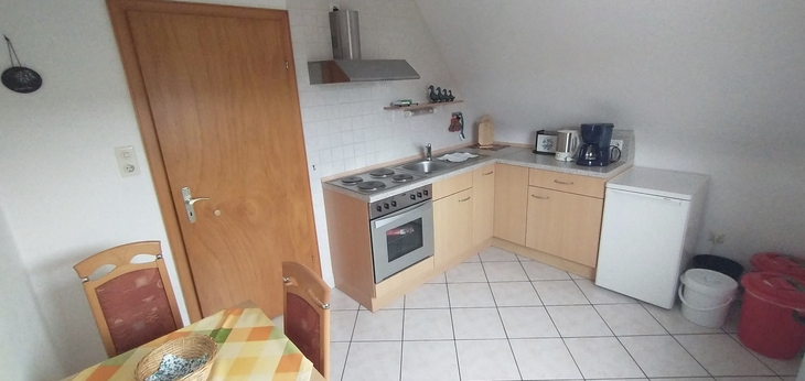 Kleine Küche mit Holzschränken, Herd, Kühlschrank und Tisch mit kariertem Tuch.