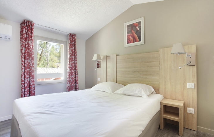 Helles Schlafzimmer mit Doppelbett, Fenster und roten Vorhängen.