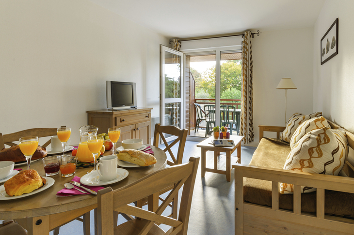 Wohnzimmer mit Frühstückstisch, Sofa, TV und offenem Balkon.