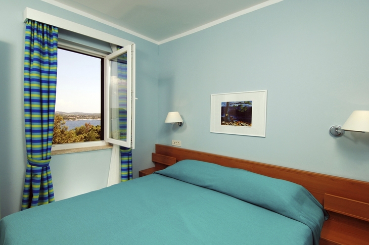 Schlafzimmer mit offenem Fenster, Blick auf Landschaft, blau-grüne Vorhänge.
