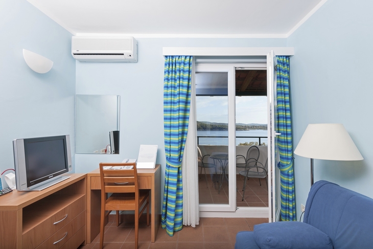 Blick in ein helles Zimmer mit Schreibtisch, TV, Sofa und Zugang zu einem Balkon mit Meerblick.