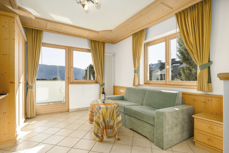 Helles Wohnzimmer mit grüner Couch, Holzmöbeln und großen Fenstern.