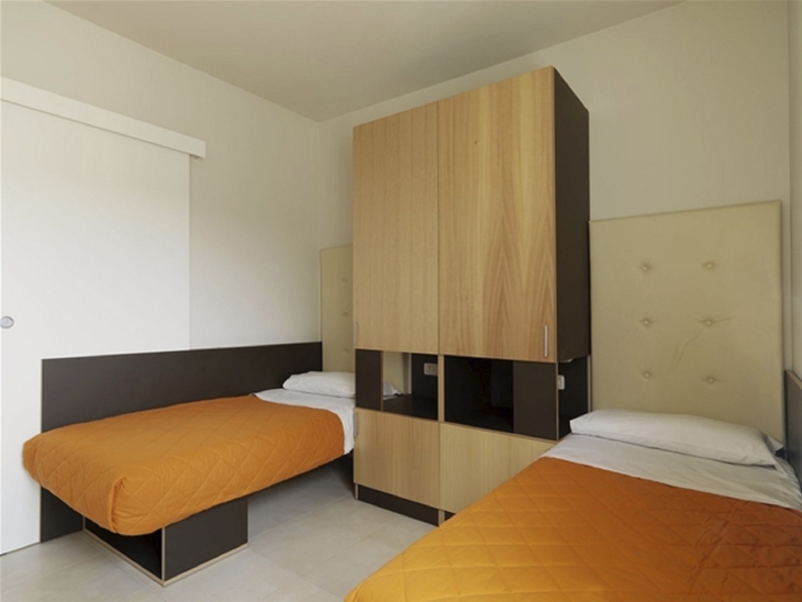 Zwei Einzelbetten mit orangefarbenen Decken in einem schlichten Schlafzimmer.