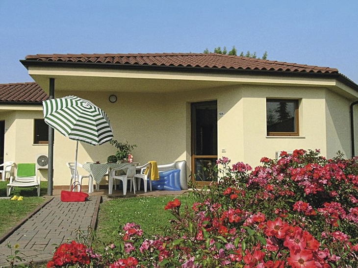 Bungalow mit Terrasse, Tisch, Stühlen und Sonnenschirm, umgeben von blühenden Rosen.