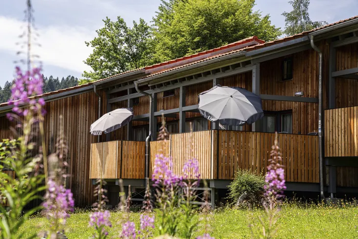 Holzgebäude mit Balkonen und Sonnenschirmen, umgeben von blühenden Pflanzen.