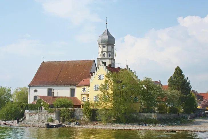 Wasserburg mit Kirchturm und Bäumen am Ufer eines Sees unter blauem Himmel mit weißen Wolken.