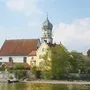Wasserburg mit Kirchturm und Bäumen am Ufer eines Sees unter blauem Himmel mit weißen Wolken.